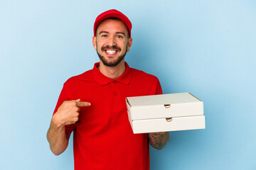 Young caucasian delivery man with tattoos holding pizzas isolated on blue background  person pointing by hand to a shirt copy space, proud and confident