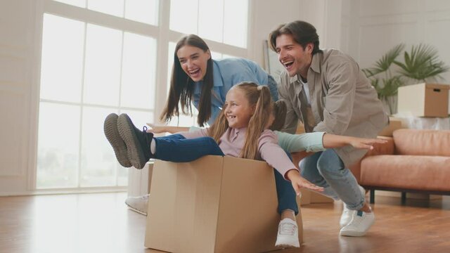 Parents pushing cardboard box with little daughters riding inside, happy family homeowners having fun on moving day
