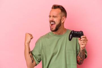Young caucasian man with tattoos holding game controller isolated on pink background  raising fist...