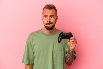 Young caucasian man with tattoos holding game controller isolated on pink background  confused, feels doubtful and unsure.