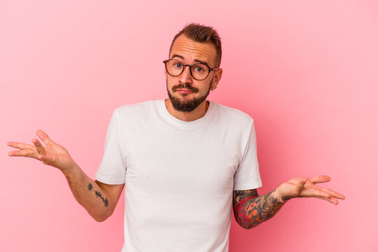 Young caucasian man with tattoos isolated on pink background  doubting and shrugging shoulders in questioning gesture.