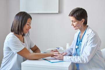 Middle-aged woman doctor in white coat measuring pulse on patient wrist, checking heartbeat of young smiling afro american female visiting private clinic, professional physician doing health check up