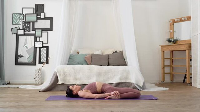 Slender, flexible, caucasian middle-aged woman with black hair in beige tracksuit performs standing asanas hero pose supta virasana on purple yoga mat in bright room with bed, a mirror, a window