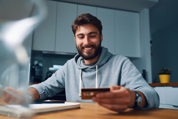Smiling young man making online payments at home with credit card and laptop