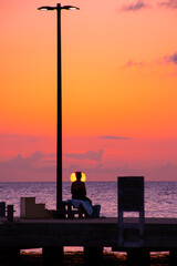 The silhouette of a young lady looking out into the Caribbean sunset. The model is sat at the end of a small dock that reaches into the sea around the Cayman Islands
