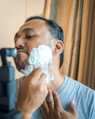 Shaving cream on the face of an Asian man. Man recording shaving process on the camera.
