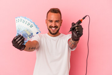 Young caucasian tattoo artist holding bills isolated on pink background