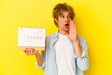 Young caucasian man with makeup holding calendar isolated on yellow background  is saying a secret hot braking news and looking aside