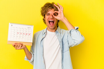 Young caucasian man with makeup holding calendar isolated on yellow background  excited keeping ok gesture on eye.