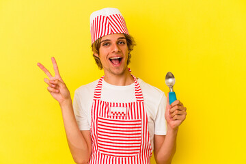 Young ice cream maker man with makeup holding spoon isolated on yellow background  joyful and carefree showing a peace symbol with fingers.