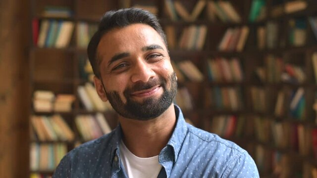 Intelligent Indian man in casual wear looks aside then at the camera with wide cheerful toothy smile, close up portrait of hispanic bearded guy standing indoor, office employee profile