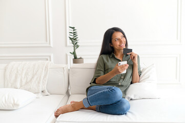 Carefree dreamy Indian woman holding credit card and smartphone, thinking about long awaited...