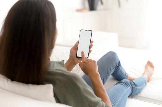 Indian young woman is using touchscreen of a smartphone, touching with a finger an empty blank screen, mockup, copy space, back view. Advertising new mobile app