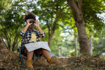 Young women teenager ethnic African American black skin wearing plaid shirt and her backpack sitting at tree base looking binocular in her hand