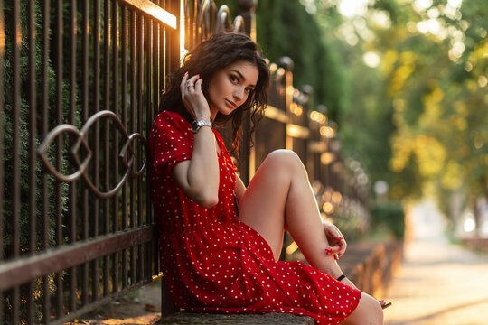 Fashion beautiful woman with curly hair in a stylish red dress sits near a fence in the city