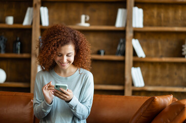 Cheerful young red-haired curly woman enjoys chatting online, sits on the couch at home and...