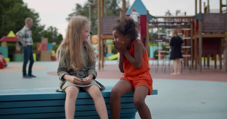 Smiling multiethnic little girls sitting on wooden bench outdoors playing