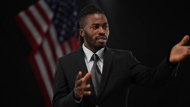 Persuasive confident African American man talking and gesturing with blurred US flag at black background. Portrait of convincing male diplomat speaking to public. Politics and governments concept