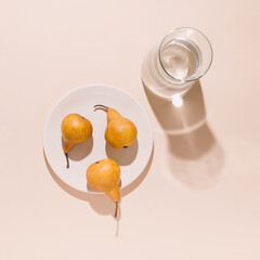 Golden yellow pears on a white ceramic plate and clear glass water pitcher casting shadows on a pale beige background. Modern autumn fruit arrangement. Cretive flat lay concept.