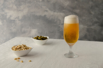 Beer glass on the table with peanut and olive snacks. White table and gray background.
