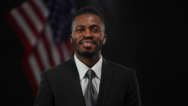 Portrait of positive confident African American man in suit talking saluting gesturing and looking at camera. Male political candidate speaking to public with American flag at black background