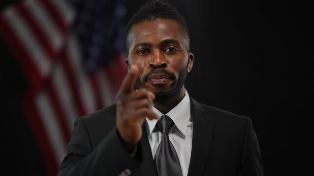 Confident African American male political candidate speaking pointing looking at camera. Front view portrait of persuasive handsome man presenting campaign with USA flag at black background