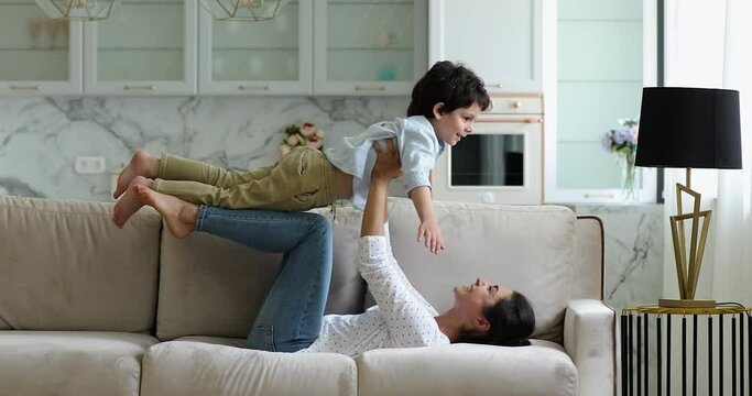 Young loving Indian ethnicity woman lying on sofa lifts on arms little mixed race son. Weekend leisure, playtime at home, dreams about family travel and holidays, active games with kids indoor concept