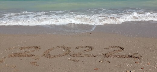 Happy New Year 2022, lettering on the beach with wave and clear blue sea. Numbers 2022 year on the sea shore, message handwritten in the golden sand on beautiful beach background. New Years concept.