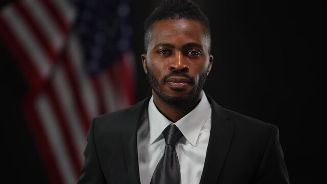 Confident African American Man Taking Off Disguise Mask Looking At Camera With Serious Facial Expression. Portrait Of Elegant Male Politician In Suit Posing At Black Background With USA Flag Behind