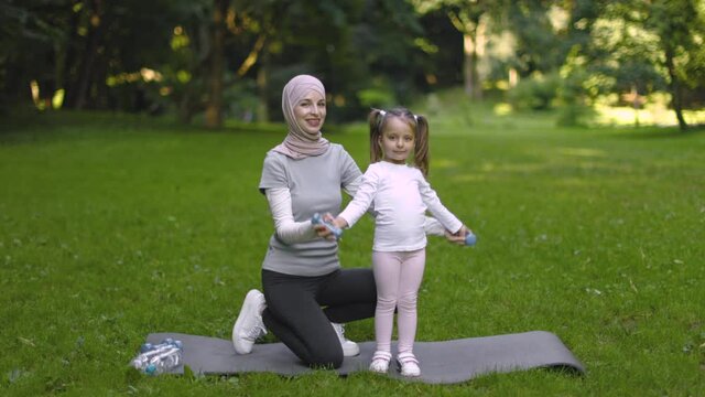Healthy lifestyle. Muslim mom in hijab helps her little daughter doing dumbbell exercises outdoors in the park. Happy kid raising dumbbells under supervision of her sporty mother