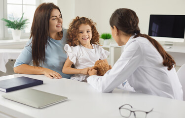 Happy mother and child seeing family doctor together. Positive female pediatrician sitting at desk in office and talking to cute cheerful little girl during her visit to clinic or hospital with mom