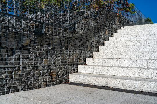 Granite staircase with fence made of stones in metal grid. Modern concrete steps with gabion fence. Modern broad stairs with stone wall and grey metallic handrail