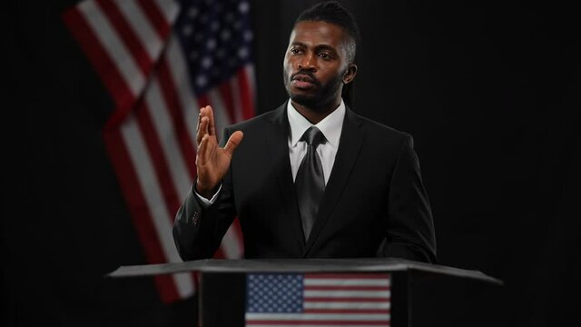 Serious professional African American politician man standing at tribune in camera flashes talking looking around. Portrait of confident diplomat answering questions with US flag at black background