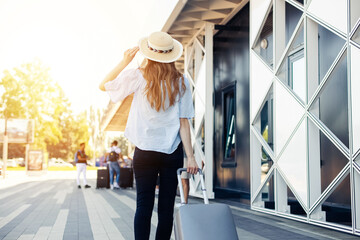 Young woman in summer clothes and hat, walks into the airport with suitcase, happy woman traveler...