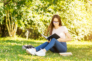 teenager is sitting on the grass, studying, a student girl under a tree in the university park with a backpack and textbooks