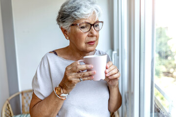 Thoughtful old woman in beige blouse drinking mug of coffee and looking through window at home. Beautiful senior woman at home standing at the window in her living room holding a cup of coffee or tea.