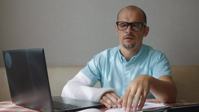 Bald man in eyeglasses with broken arm in plaster cast having stress while working remotely from home, touching his head and looking at notes, laptop standing beside. Problems in business