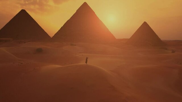 Slow motion. Silhouette of woman in traditional arab dress rises her arms at the sunset with pyramids on the background.
