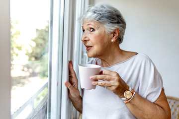 Aged woman at home standing near the window drinking tea looking out the window dreaming. Senior lady having coffee and looking thoughtfully out of window. Shot of an attractive old woman drinking tea