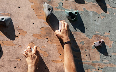 Close-up photo of male hands gripping climbing holds on worn wall outside. Man doing climbing...