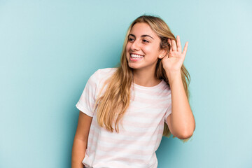 Young caucasian blonde woman isolated on blue background  trying to listening a gossip.