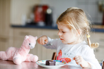 Cute funny toddler girl eating chocolate ice cream at home. Happy healthy baby child feeding plush...