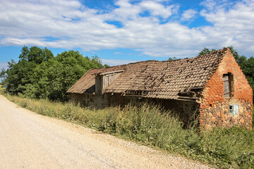 An old dilapidated stone and red brick shed with a tiled roof, standing by the road.