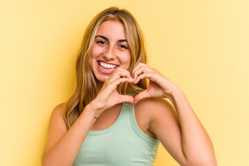 Young caucasian blonde woman isolated on yellow background  smiling and showing a heart shape with hands.