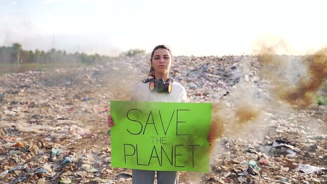 young woman activist with a poster in hands save the planet stands middle a landfill with a respirator. black smoke. female student girl protests against environmental pollution nature conservation