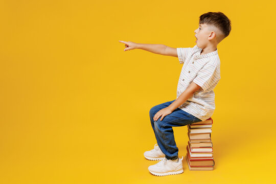 Side View Young School Boy 5-6 Years Old In Casual Clothes Sit On Pile Of Book Point Finger On Workspace Isolated On Plain Yellow Background Studio Childhood Children Kids Education Lifestyle Concept