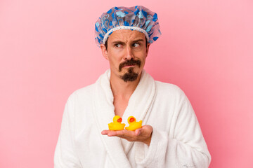Young caucasian man going to the shower with rubber ducks isolated on pink background confused, feels doubtful and unsure.