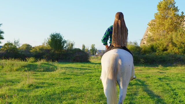 A teenage country girl riding a horse on a green field on a sunny day. A young cowgirl astride a white horse, back view