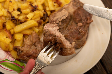 Someone is cutting a piece of juicy roast meat on a plate of fried potatoes with a knife and fork. Close-up Selective focus.