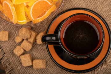 Multicolored shaped marmalade lemon slices in a glass vase, pieces of brown cane sugar and a mug of coffee, close-up, selective focus.